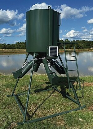 A FeedVault unit installed on a green gravity-fed protein feeder standing on grass beside a pond. A solar panel is mounted on the side of the hopper, an access ladder runs up the right leg, and the linear actuators that open and close the feed tubes are fitted beneath the hopper.
